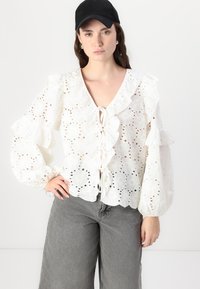 White eyelet blouse with ruffled sleeves and a V-neck, featuring a lace-up front. Paired with gray wide-leg jeans and a black cap.