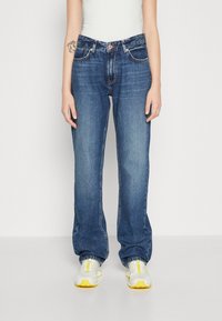 Vaqueros de mezclilla azul oscuro con corte recto, diseño de cinco bolsillos, costuras naranjas y un cierre clásico de botón, combinados con zapatillas grises.