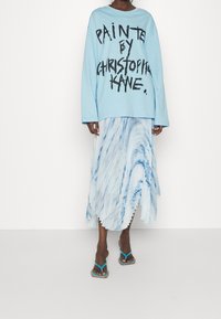 Hellblaue, oversized Sweatshirt mit schwarzem Text, kombiniert mit einem plissierten, midi-langen Rock in weiß-blauem Marmor-Muster und türkisen Flip-Flops.