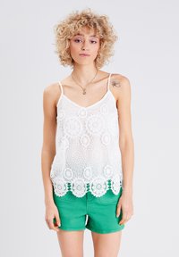 Camisole en dentelle blanche avec des motifs en crochet circulaires complexes, dotée de bretelles réglables et d'un ourlet festonné, portée sur des shorts verts.