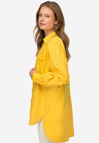 Camicia gialla con bottoni, maniche lunghe, due tasche frontali, orlo curvo e dettagli ricamati verdi sul retro. Presenta una vestibilità comoda.