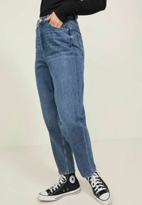 Hochtaillierte blaue Jeans aus Denim mit lockerer Passform, ausgestattet mit einem klassischen Fünf-Taschen-Design und einem Knopfverschluss. Kombiniert mit schwarzen High-Top-Sneakers.