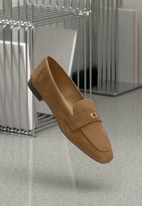 Mocassino in suede marrone con tacco piatto, caratterizzato da un dettaglio a emblema dorato sulla fascia. Texture liscia e forma classica.