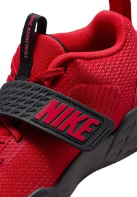 Punainen urheilukenkä, jossa on kuvioitu päälliosa, säädettävä hihna mustilla metalliosilla ja näkyvä Nike-brändäys punaisena mustalla pohjalla.