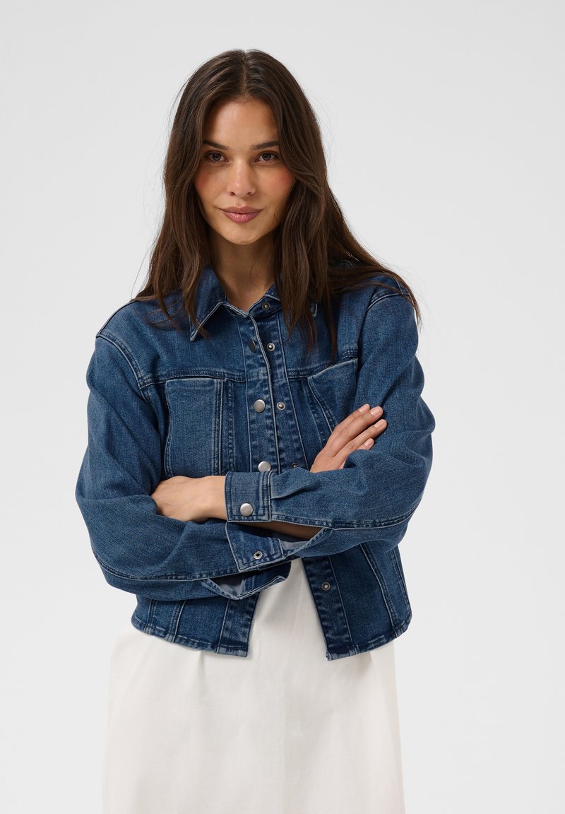 Femme aux longs cheveux bruns portant une veste en jean bleue sur une robe blanche, debout les bras croisés devant un fond uni.