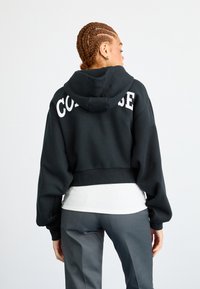 Black cropped hoodie with white "CONVERSE" text on the back. Features a hood and wide, slightly puffed sleeves. Worn over a white ribbed top.