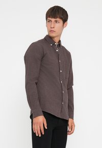 Brun langermet skjorte med knapper laget av teksturert stoff, med spiss krage, en brystlomme og buet nederkant.