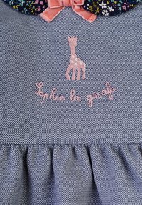 Blauer Denim-Stoff mit einem rüschengesäumten Saum, versehen mit einem pinken Giraffen-Logo und dem Text "Sophie la girafe" in pinker Stickerei. Blütengemusterter Kragen.
