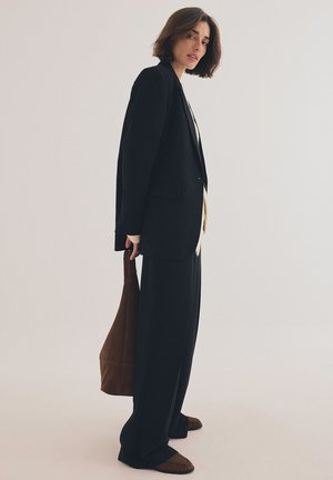 Schwarze, oversized Blazerjacke und weit geschnittene Hose, kombiniert mit einem cremefarbenen Top. Hält eine braune Wildleder-Tote-Tasche. Weiche Textur und klare Linien.