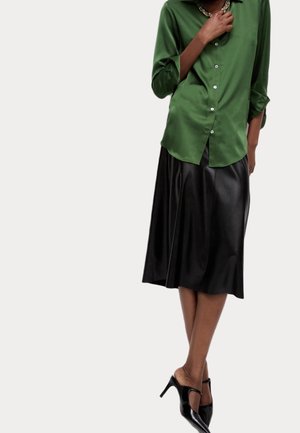 Osoba nosící zelenou saténovou košili na knoflíky, černou sukni po kolena, zlatý řetízkový náhrdelník a černé lodičky s pásky kolem kotníků.