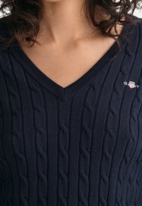 Vrouw in een marineblauwe kabelgebreide V-halstrui met een klein geborduurd GANT-logo op de linkerkant van de borst.