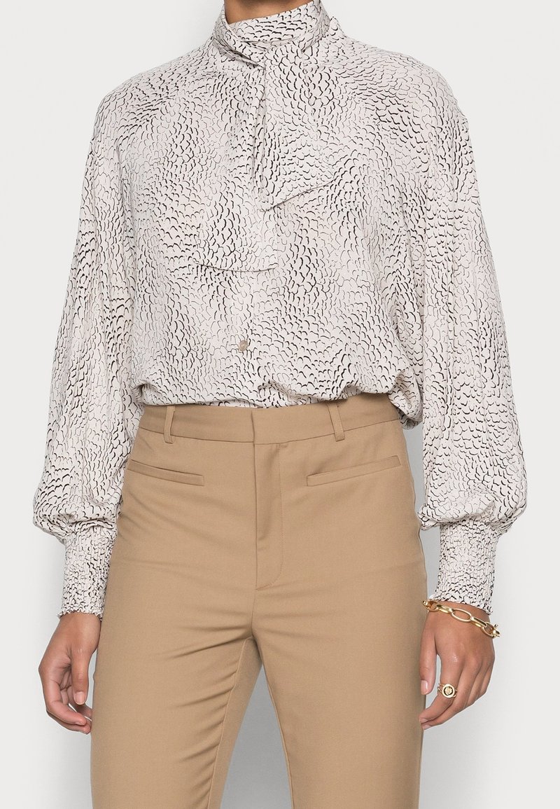 Blusa a maniche lunghe in beige chiaro con motivo astratto nero, caratterizzata da un alto colletto e dettagli arricciati, abbinata a pantaloni color sabbia.