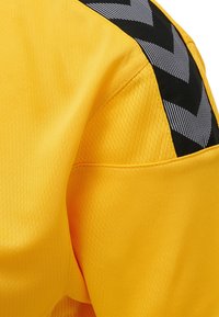 Gul idrottströja med fuktavledande meshmaterial och svarta chevrondetaljer längs axeln. Slät yta, design med rund hals.