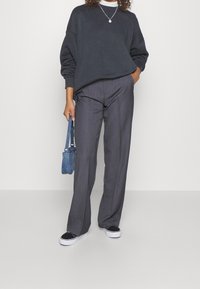 Felpa oversize grigia scura abbinata a pantaloni leggeri grigi a pieghe, accentuati da una piccola borsa blu e sneakers nere con suole bianche.