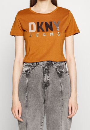 Kvinde iført en brun DKNY Jeans t-shirt og grå højttaljede jeans, vist fra skuldre til hofter, stående med afslappede hænder.