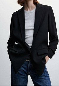 Blazer nero su misura con design a un petto, rever a scasso e tasche frontali, abbinato a una camicia grigia a costine e jeans blu.