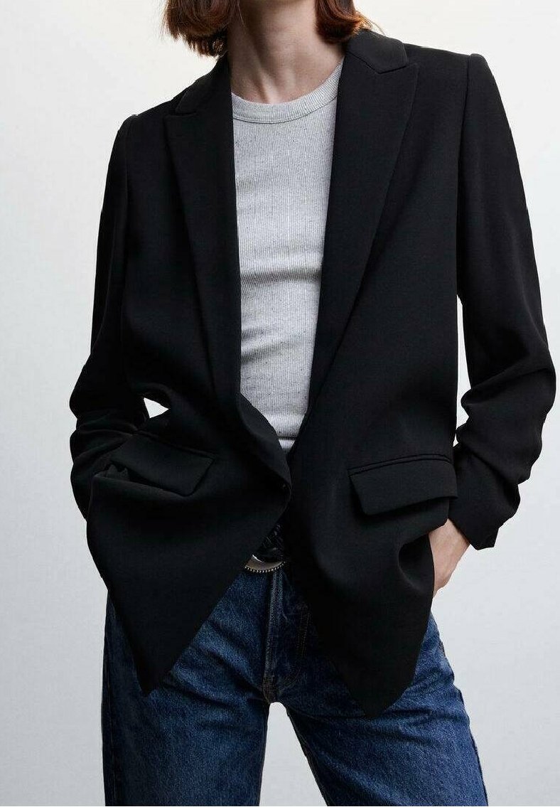 Blazer nero su misura con design a un petto, rever a scasso e tasche frontali, abbinato a una camicia grigia a costine e jeans blu.