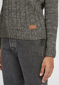 Szary sweter dzianinowy z textured kabelkowym wzorem, z skórzaną metką z nazwą marki. Noszony z ciemnoszarymi dżinsami.