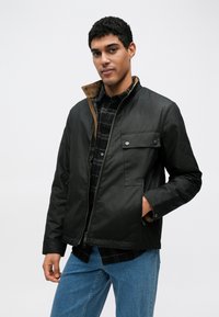 Jachetă neagră cu fermoar și închidere cu capse, guler maro contrastant și buzunar frontal; purtată peste o cămașă în carouri și blugi albaștri.