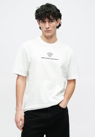 Άσπρο βαμβακερό μπλουζάκι με εκτυπωμένο μαύρο λογότυπο Μέδουσας και κείμενο μπροστά. Κοντά μανίκια και χαλαρή εφαρμογή, συνδυασμένο με μαύρο παντελόνι.