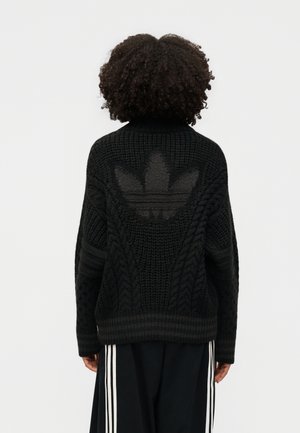 Persona con cabello rizado de espaldas, vistiendo un suéter negro de lana con trenzas y un gran logo texturizado en la espalda, y pantalones negros con rayas blancas.