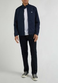 Giacca leggera navy con colletto rialzato, logo bianco sul petto, abbinata a una maglietta bianca, jeans scuri e sneakers nere.