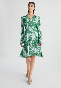 Vestito camicia verde e bianco a maniche lunghe con motivo a foglie, vita con cintura, chiusura frontale con bottoni e orlo svasato. Abbinato a scarpe testurizzate.