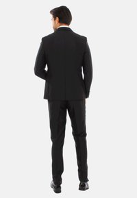 Completo nero con una vestibilità sartoriale, tessuto liscio e pantaloni slim-fit. La vista posteriore mostra uno spacco singolo e dettagli minimi.