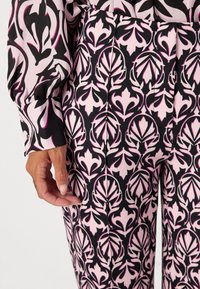 Die Hose zeigt ein schwarz-rosanes und weißes Blumenmuster mit kräftigen Konturen und besteht aus einem glatten Stoff. Eine sichtbare Hand ist in der Nähe des Bunds platziert.