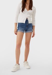 White ribbed cardigan with button front, paired with denim shorts featuring rolled hems. Notable white sneakers complete the outfit.