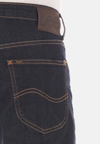 Donkerblauwe denim jeans met zichtbare oranje stiksels, voorzien van een leren taillelabel en een achterzakontwerp met ronde hoeken.