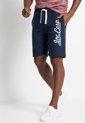 Shorts en coton bleu marine avec une taille à cordon et un texte graphique blanc sur la jambe gauche. Assortis avec des chaussures de sport blanches.