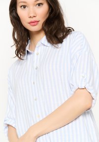 Šviesiai mėlynas ir baltos juostelės marškiniai su mygtukų apykakle, suktomis rankovėmis ir baltais mygtukais, pagaminti iš minkštos, lengvos medžiagos.