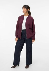 Burgunderfarbene Bomberjacke mit einem Frontreißverschluss, kombiniert mit hochgeschnittenen, weit geschnittenen dunkelblauen Jeans und braunen Stiefeletten mit Absatz.