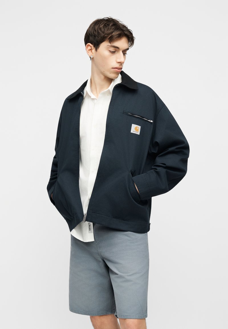 Jonge man met een zwart jack met ritssluiting over een wit shirt en grijze korte broek, staand met de handen in de zakken van het jack, kijkend naar de zijkant.