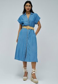 Svetlo modra denim obleka s kratkimi rokavi, sprednjim zapenjanjem na gumbe, pasom, ki ga lahko povežemo, ter obleko srednje dolžine s sprednjim razporkom. Oblečena z espadrilami na visoki peti.