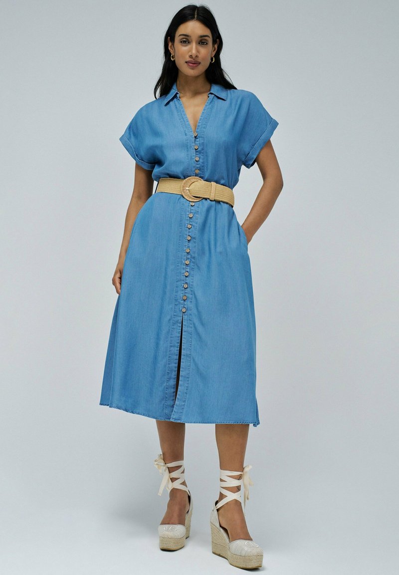 Svetlo modra denim obleka s kratkimi rokavi, sprednjim zapenjanjem na gumbe, pasom, ki ga lahko povežemo, ter obleko srednje dolžine s sprednjim razporkom. Oblečena z espadrilami na visoki peti.