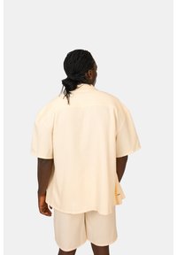 Camicetta corta beige chiaro a maniche corte con righe verticali e orlo curvo, abbinata a pantaloni corti leggeri coordinati. Tessuto semplice e testurizzato.