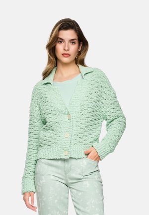 Woman wearing a light green textured buttoned cardigan, matching floral pants, and a plain light green top, posing with one hand in pocket.