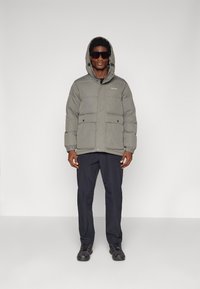 Puffer jacket in olive green, featuring a hood, two front pockets, and elastic cuffs. Paired with dark trousers and black sneakers.