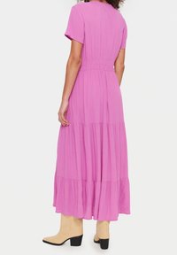 Maxi šaty v sytě růžové barvě, krátké rukávy, těsně se přizpůsobující pas a design sukně ve vrstvách. Nosené s béžovými kozačkami na podpatku. Hladká textura.