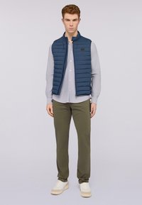 Chaleco acolchado azul marino con patrón de acolchado horizontal, que se lleva sobre una camiseta gris claro, combinado con pantalones verde oliva y zapatillas blancas.