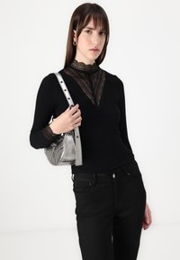 Čierne dlhé rukávy s detailom čipkového výstrihu, v kombinácii s čiernymi džínsami. Model drží metalickú striebornú tašku s popruhom cez rameno.
