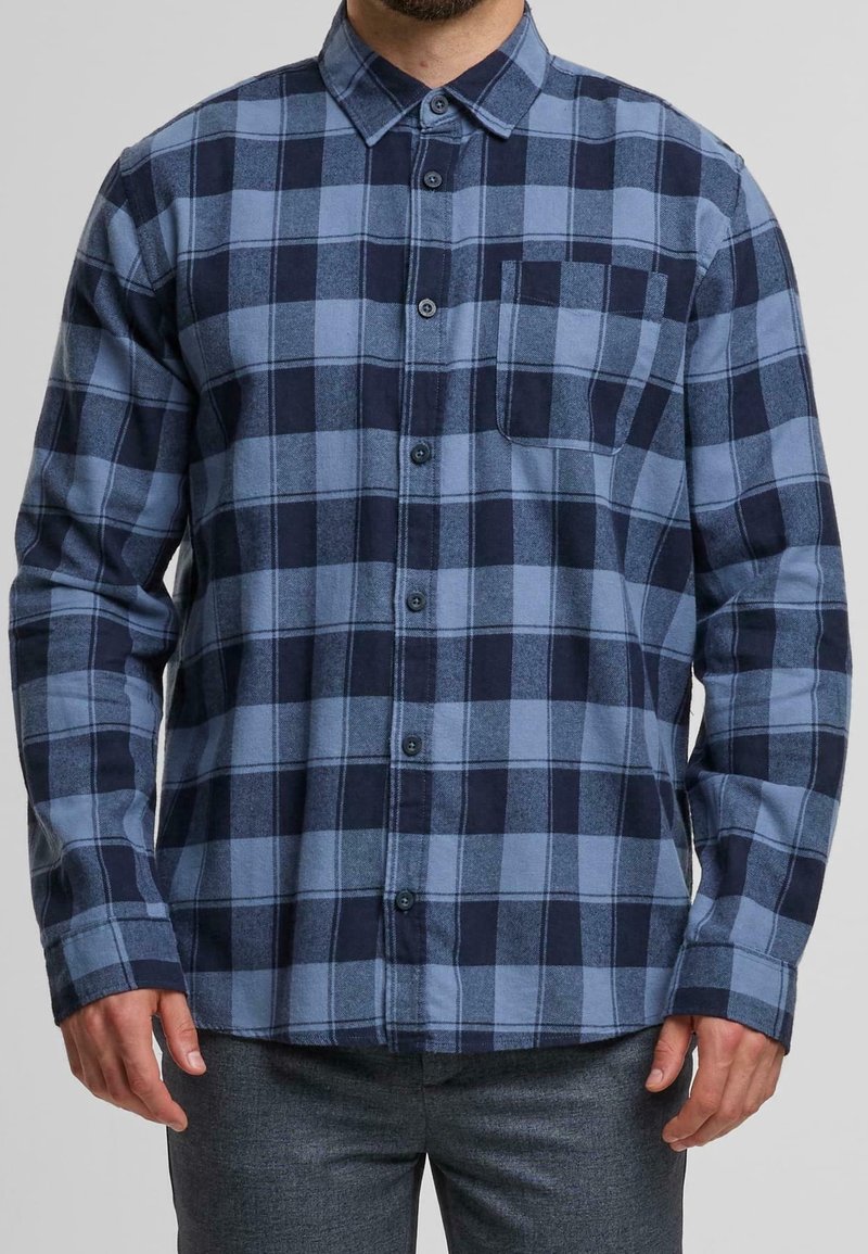 Homme portant une chemise à carreaux bleue et bleu foncé à manches longues avec poche poitrine et pantalon gris.