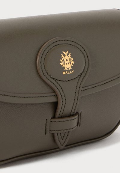 Sac en cuir vert foncé avec un rabat arrondi, présentant un logo circulaire en or et des coutures décoratives. Design épuré et texture douce.
