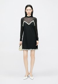 Melna kleita ar viļņotu, izrotātu apakšu, pārklāta ar caurspīdīgu melnu garu piedurkņu blūzi. Gluda auduma, strukturēta forma un vienkārši aksesuāri.