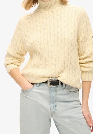 Person iført en cremefarvet kabelstrikket rullekravepullover med opkørte ærmer og lyseblå højttaljede jeans med sort bælte.