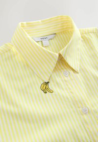 Gelb gestreiftes Hemd mit klassischem Kragen, weißen Knöpfen und einem kleinen gestickten Bananenmotiv auf der linken Seite.