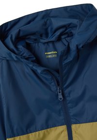 Giubbotto antivento blu con una parte inferiore gialla, dotato di zip frontale, cappuccio e tessuto liscio e lucido. Etichetta visibile all'interno.