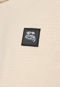 Tissu en tricot de couleur crème avec un motif texturé, présentant une petite étiquette noire avec un design graphique et un texte en blanc.